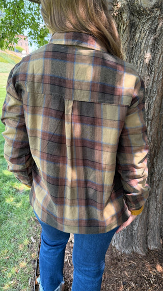 A person viewed from behind wearing a long-sleeve plaid flannel shirt in beige, brown, and muted blue, paired with blue jeans, standing outdoors next to a tree trunk.