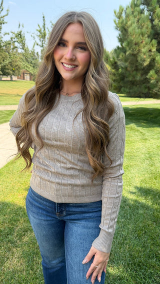 A smiling woman with long wavy light brown hair stands outdoors on a grassy lawn wearing a beige cable-knit long-sleeve sweater and blue jeans.