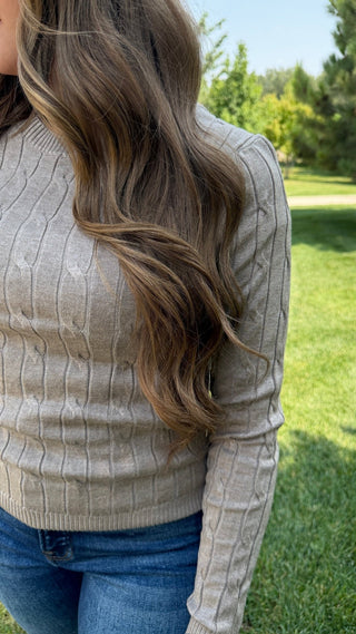 A woman wearing a beige cable-knit long-sleeve sweater and blue jeans, standing outdoors on a grassy lawn with trees in the background.