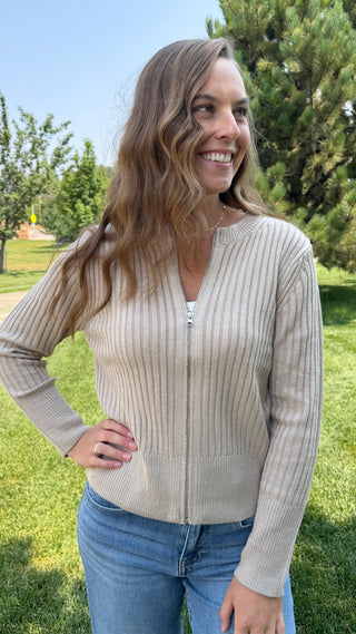 A woman wearing a beige ribbed zip-up sweater and blue jeans, standing outdoors on a grassy lawn with trees in the background.
