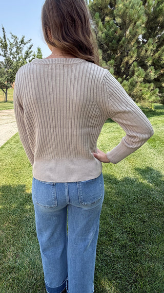 A woman viewed from behind wearing a beige ribbed knit sweater and light blue jeans, standing outdoors on a grassy lawn with trees in the background.