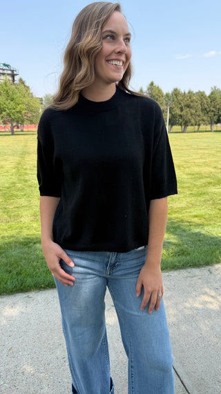A woman wearing a black short-sleeve mock neck t-shirt and light blue jeans, standing outdoors on a paved path with a grassy park and trees in the background.