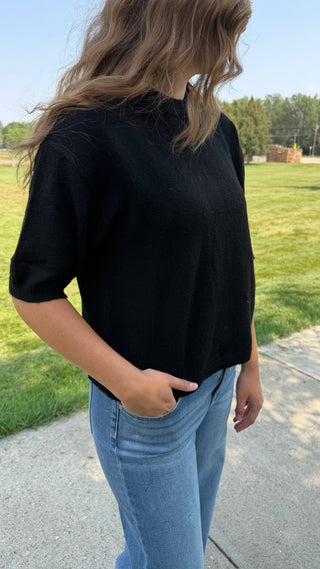 A woman wearing a black short-sleeve knit top and light blue jeans, standing outdoors on a paved path with a grassy field and trees in the background.