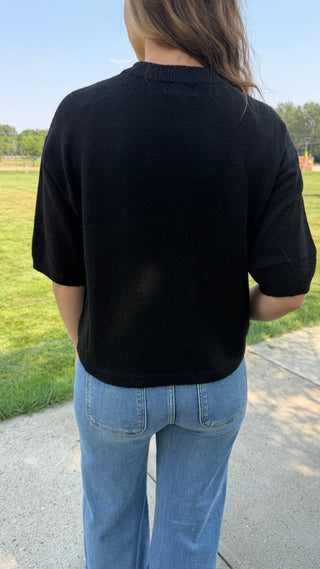 A person viewed from behind wearing a black short-sleeve knit top and light blue jeans, standing outdoors on a paved path with grass and trees in the background.