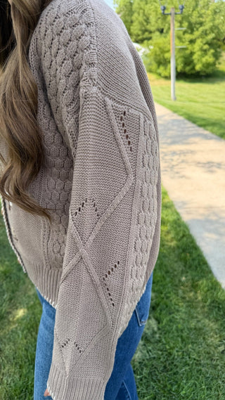 A person outdoors wearing a taupe cable-knit sweater and blue jeans, with grass, trees, and a paved path in the background.