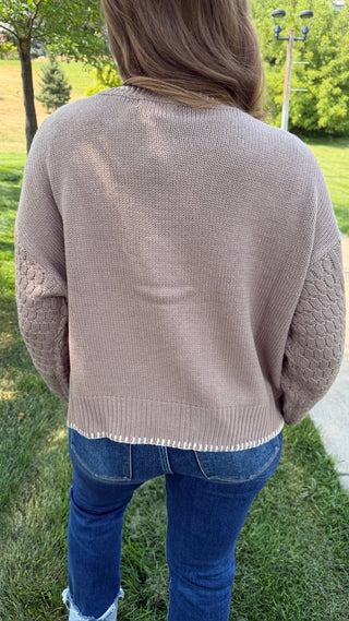 A person viewed from behind wearing a light taupe knit sweater with textured cable-knit elbow patches and blue jeans, standing outdoors on a grassy lawn.