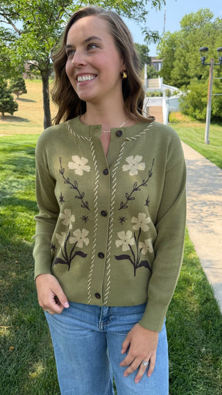 A woman wearing an olive green knit cardigan with cream and brown floral embroidery, paired with blue jeans, standing outdoors on a grassy lawn next to a paved path.