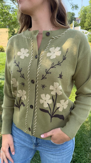 A woman wearing an olive green cardigan with cream and dark brown floral knit patterns, paired with blue jeans, photographed outdoors on a grassy lawn.