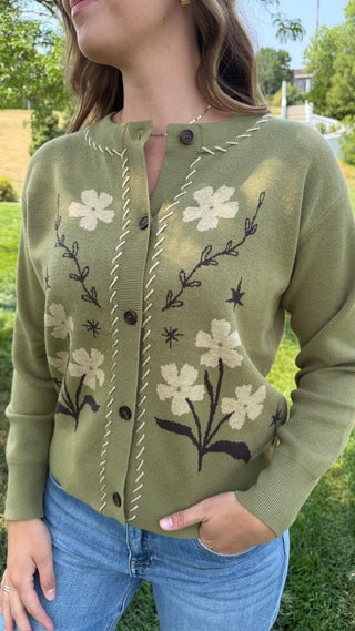 A woman wearing an olive green knit cardigan with cream and dark brown floral embroidery, paired with blue jeans, photographed outdoors on a grassy lawn.