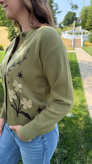 A woman wearing an olive green knit cardigan with cream and black floral embroidery, paired with blue jeans, standing outdoors on a grassy lawn.