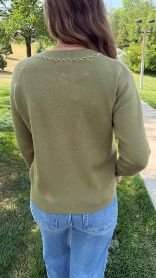 A woman viewed from behind wearing an olive green long-sleeve knit sweater with decorative stitching across the upper back, paired with light blue jeans, standing outdoors on a grassy lawn.