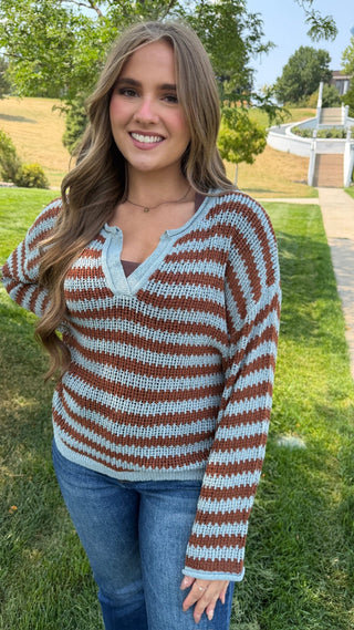 A woman wearing a brown and light blue striped knit sweater with a split V-neck, standing outdoors on a grassy lawn with trees and a pathway in the background.