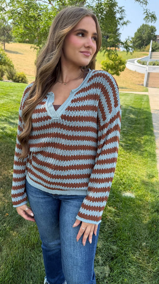 A woman wearing a brown and light blue striped knit sweater with a V-neck, standing outdoors on a grassy lawn.