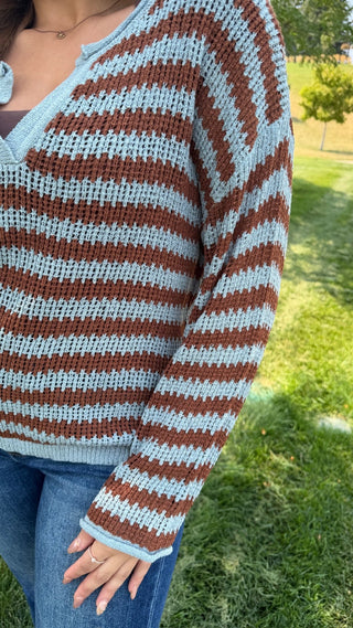 A person outdoors wearing a chunky knit cardigan with horizontal brown and light blue stripes, paired with blue jeans. The background shows green grass and trees.