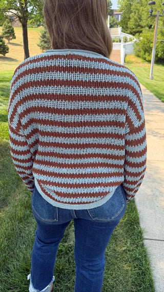 A person viewed from behind wearing a cropped knit sweater with horizontal rust brown and light gray stripes, paired with blue jeans, standing outdoors on a grassy lawn next to a paved walkway.