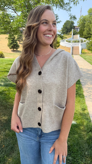A woman smiling outdoors wearing a light beige knit short-sleeve button-front cardigan with dark buttons and side pockets, paired with blue jeans.