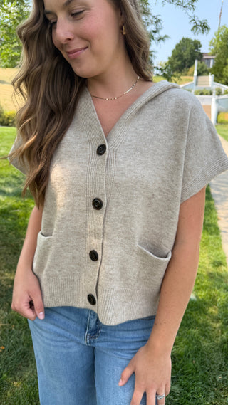 A woman wearing a light beige knit short-sleeve button-front cardigan with dark buttons and front patch pockets, paired with blue jeans, standing outdoors on a grassy lawn.