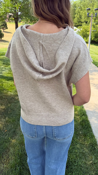 A woman viewed from behind wearing a light beige knit hooded top with short sleeves and blue jeans, standing outdoors on grass near a walkway.