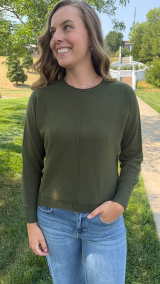 A woman wearing an olive green long-sleeve knit sweater and light blue jeans, standing outdoors on a grassy lawn next to a paved path.