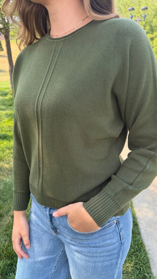 A woman wearing an olive green knit sweater with a crew neck and ribbed cuffs, paired with light blue jeans, photographed outdoors on a grassy lawn.