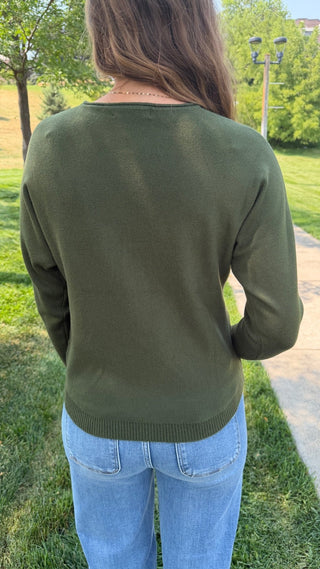 A woman viewed from behind wearing an olive green long-sleeve knit sweater and light blue jeans, standing outdoors on a grassy lawn next to a paved walkway.