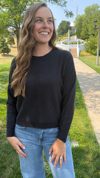 A woman smiling outdoors wearing a black long-sleeve knit sweater and light blue jeans, standing on a grassy path with trees and a walkway in the background.