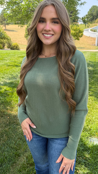 A woman with long wavy brown hair stands outdoors on a grassy lawn wearing a sage green long-sleeve waffle-knit sweater and blue jeans.