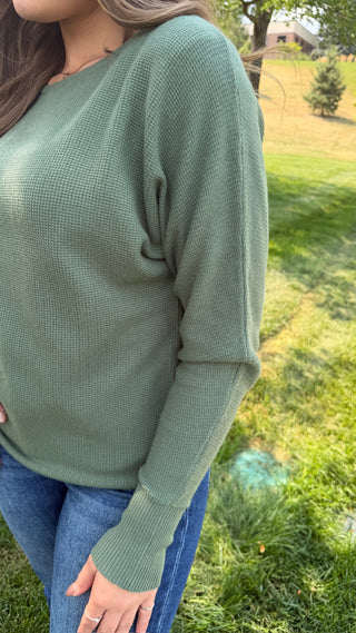 A woman wearing a sage green waffle-knit long-sleeve sweater and blue jeans, standing outdoors on a grassy lawn.