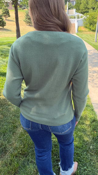 A woman viewed from behind wearing an olive green waffle-knit long-sleeve sweater and blue jeans, standing outdoors on a grassy lawn with trees and a path in the background.