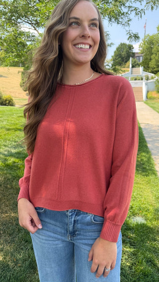 A woman wearing a rust-colored knit sweater and blue jeans, standing outdoors in a grassy park area with trees and a pathway in the background.