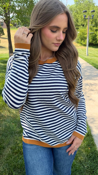 A woman wearing a navy and white horizontally striped long-sleeve top with orange ribbed trim, paired with blue jeans, standing outdoors on a paved path in a park.