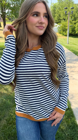 A woman wearing a navy and white horizontally striped long-sleeve top with mustard yellow ribbed trim, paired with blue jeans, standing outdoors on a grassy lawn next to a paved path.