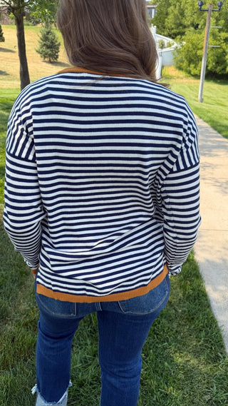 A person viewed from behind wearing a navy and white horizontally striped long-sleeve top with a mustard yellow hem, paired with blue jeans, standing outdoors on a grassy lawn next to a paved path.