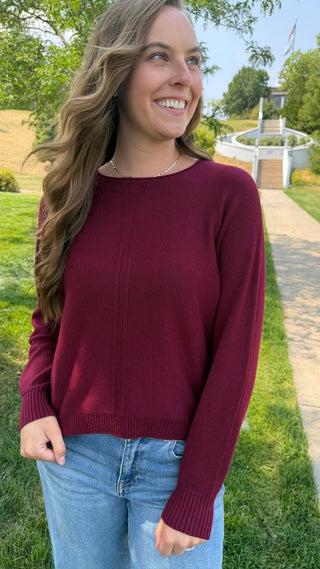 A woman wearing a long-sleeve crewneck sweater in burgundy, paired with light blue jeans, standing outdoors on a grassy lawn with a pathway and trees in the background.