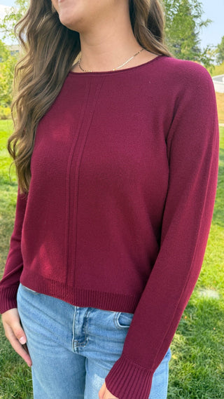 A woman wearing a long-sleeve crewneck sweater in burgundy, paired with light blue jeans, photographed outdoors on a grassy lawn.