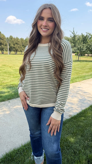 A woman standing outdoors on a paved path in a grassy park, wearing a cream and olive green horizontally striped long-sleeve knit top with blue jeans.