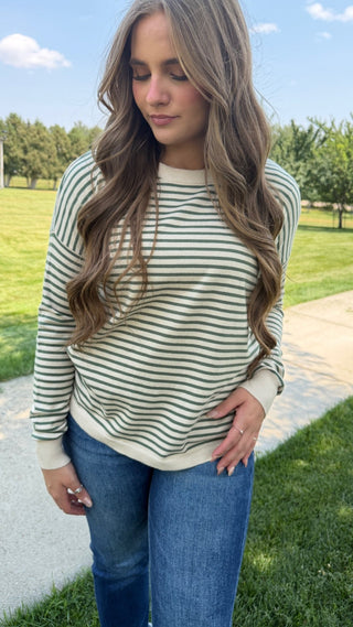 A woman wearing a cream and dark green striped long-sleeve crewneck sweatshirt with blue jeans, standing outdoors on a paved path with a grassy park in the background.