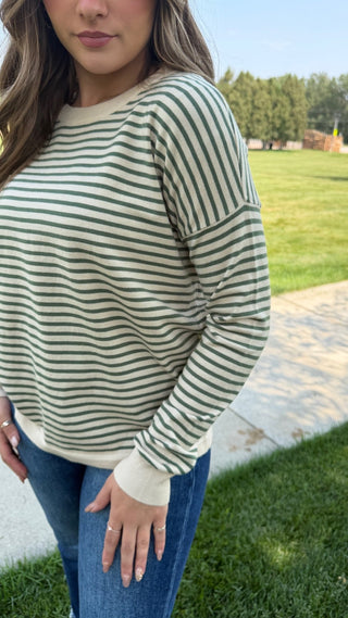 A woman wearing a long-sleeve crewneck top with horizontal stripes in sage green and cream, paired with blue jeans, standing outdoors on a paved path with a grassy lawn in the background.
