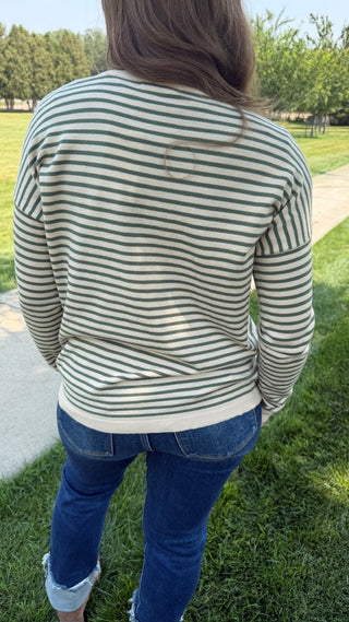 A person viewed from behind wearing a long-sleeve striped top with blue jeans, standing outdoors on grass near a paved path.