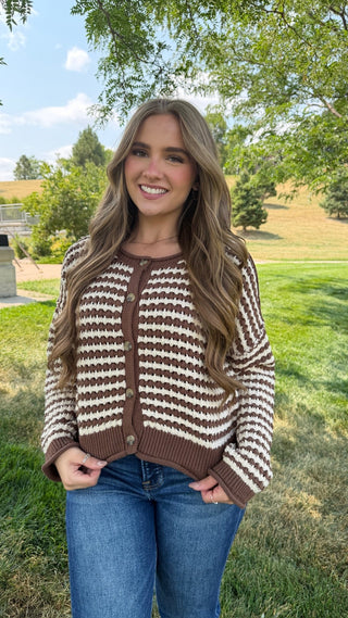 A woman wearing a brown and cream striped knit cardigan sweater and blue jeans, standing outdoors in a grassy park area with trees and hills in the background.