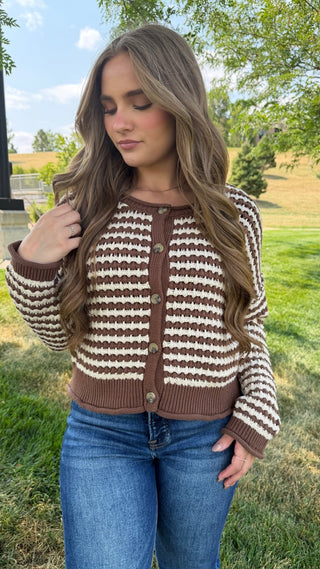 A woman wearing a brown and cream striped knit cardigan with blue jeans, standing outdoors in a grassy park area.