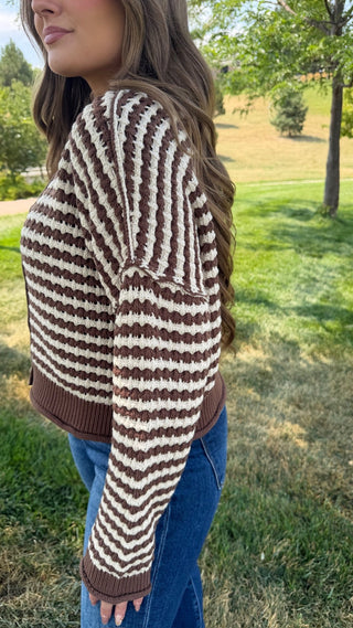 A woman wearing a brown and cream striped knit cardigan sweater with blue jeans, standing outdoors in a grassy park area.