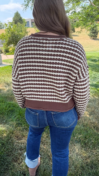 A person viewed from behind wearing a brown and cream striped knit sweater and blue jeans, standing outdoors on a grassy lawn with trees in the background.