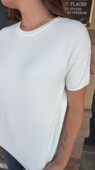 A close-up of a woman wearing a white ribbed short-sleeve crewneck top, showing the texture of the fabric and her arm.