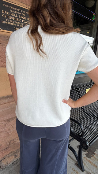 A woman viewed from behind wearing a white short-sleeve ribbed knit top and navy blue pants, standing outdoors next to a brick wall and a bench.
