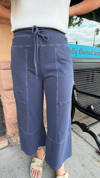 A person wearing slate blue wide-leg cropped pants with a drawstring waist, paired with a white ribbed top and beige sandals, standing outdoors near a bench.