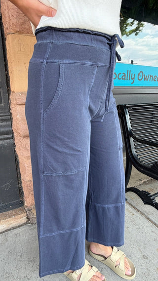 A person wearing wide-leg cropped pants in a dusty slate blue color, styled with a white top and beige slide sandals, standing outdoors near a storefront.