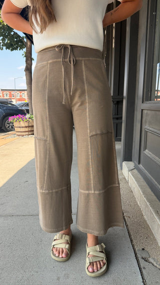 A person standing outdoors on a sidewalk wearing loose, wide-leg taupe pants with a drawstring waist, paired with a white top and beige slide sandals.