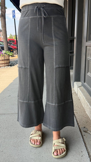 A person wearing dark gray wide-leg cropped pants with a drawstring waist and light-colored slide sandals, standing on a sidewalk outdoors.