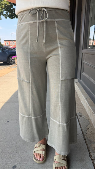A person wearing light beige wide-leg cropped pants with white contrast stitching and a drawstring waist, paired with beige slide sandals on a sidewalk.